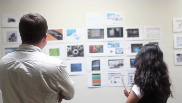 Two workshop participants stand in front of a wall, and they are looking at a collection of printouts that have been taped to the wall. Each printout features colorful images of products, UIs, graphics, and textures